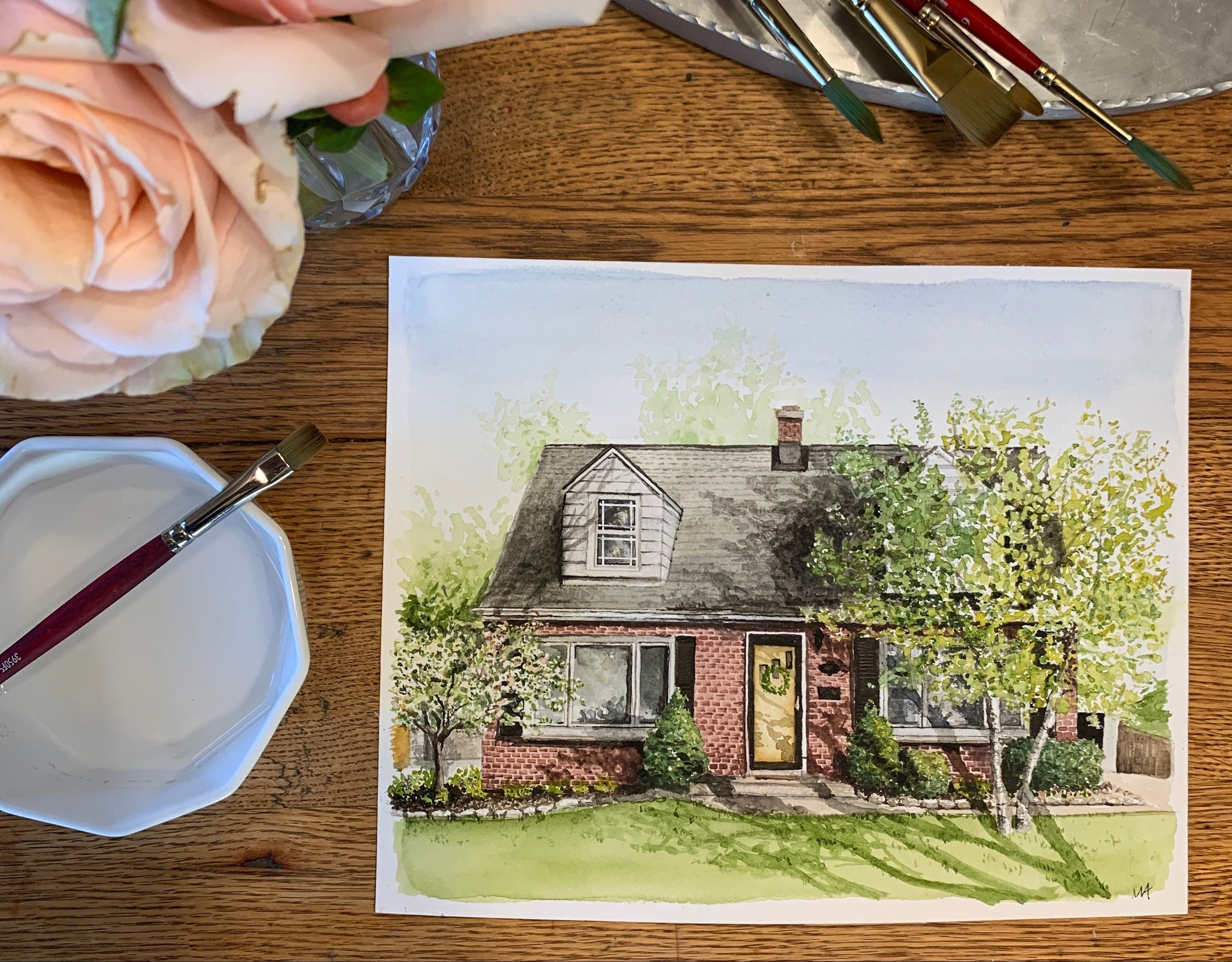Painting of a house on paper with art supplies including brushes and flowers on a wooden table.
