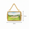 A gold-tone rectangular hanging clutch or pouch with a small landscape painting inset showing a baseball field, overlaid with dimension arrows and labels reading 3 in wide and 2 in tall on a white background.