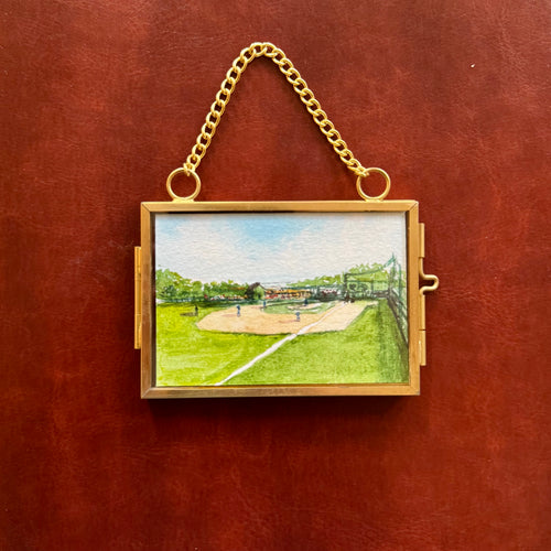 A gold-tone hanging locket box featuring a hand-painted watercolor baseball field scene, set against a textured reddish-brown leather background.